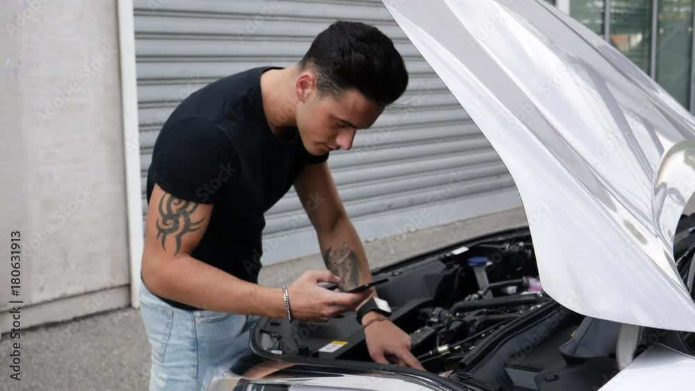 Young man standing near his car and trying to understand how to fix it with online advice found on cellphone Young man standing near his car and trying to understand how to fix it with online advice found on cellphone