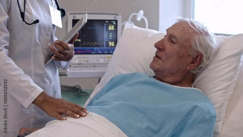 Elderly man lying on bed in comfortable hospital ward and speaking with female doctor standing with digital tablet Elderly man lying on bed in comfortable hospital ward and speaking with female doctor standing with digital tablet