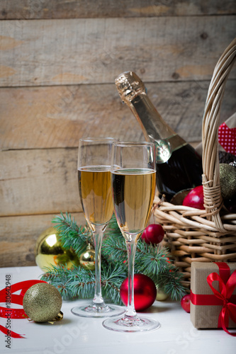 Two glasses of champagne with christmas basket with bottle, gifts with red satin ribbon, candy canes, pine cones, golden garlands on dark brown wooden background, new year celebration concept