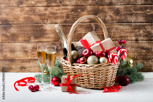 Two glasses of champagne with christmas basket with bottle, gifts with red satin ribbon, candy canes, pine cones, golden garlands on dark brown wooden background, new year celebration concept