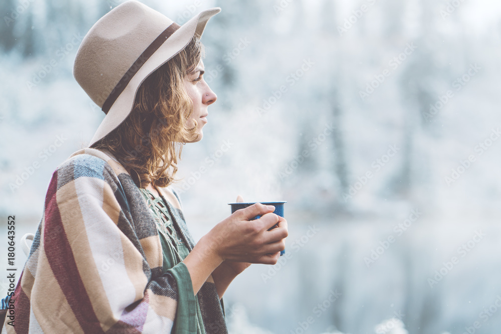 Fototapeta premium Pretty woman with hot drink in hands spending vacations among stunning winter landscape. Traveling in mountains wilderness. Wanderlust and boho style