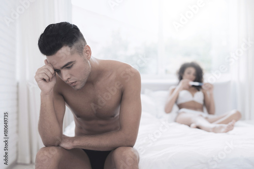 The girl is holding a pregnancy test and frowning. She is not happy that she showed the test. She sits on a white bed in her underwear. A young man is sitting by her side. He frowns