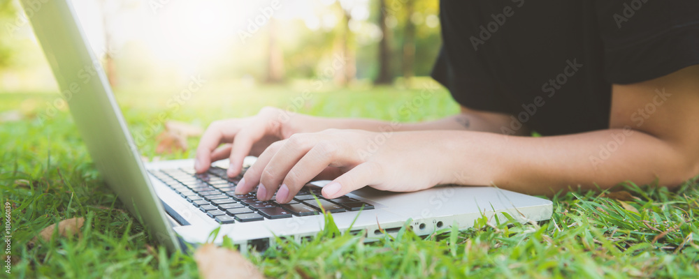 Young asian woman's legs on the green grass with open laptop. Girl's hands on keyboard. Distance learning concept. Happy hipster young asian woman working on laptop in park. Panoramic banner. - obrazy, fototapety, plakaty 