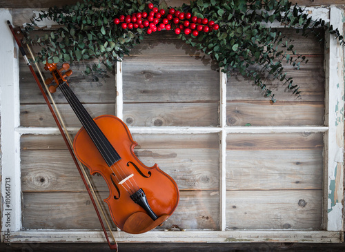 horizontal image of a violin lying on a rustic wood surface with a white window frame and green holly with red berries. © nat2851terry