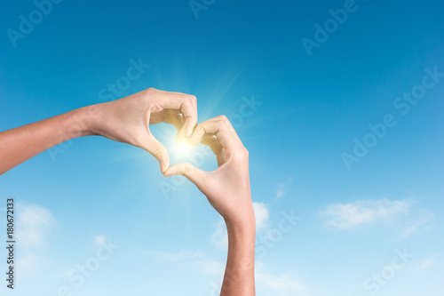 hands making a heart shape in the blue sky with sunlight