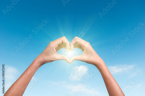 hands making a heart shape in the blue sky with sunlight