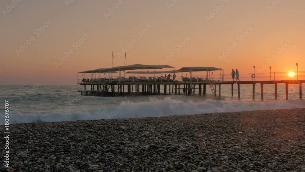 Colorful sunset. Sea view with waves and foam, pier and pebble beach. Silhouettes of happy floating people in the sea. Colorful sunset. Sea view with waves and foam, pier and pebble beach. Silhouettes of happy floating people in the sea.