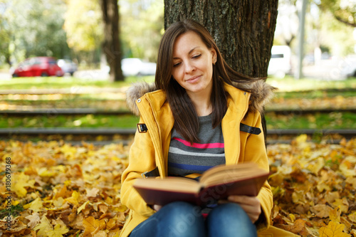 A beautiful happy smiling brown-haired woman in yellow coat and jeans sitting under the maple tree with a red book in fall city park on a warm day. Autumn golden leaves. Reading concept
