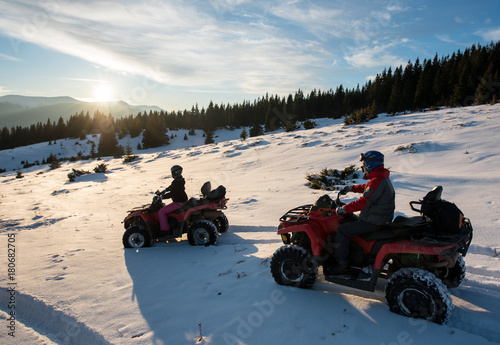 Young male and female riders sitting on quad bikes on snow, enjoying sunset in the the mountains in winter