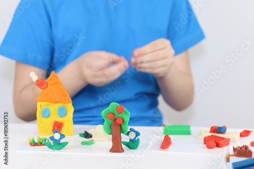Child hands molding bright house, sheep, flowers from plasticine on table in room