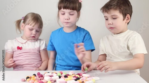 Three happy children eat candies and sit at table in white studio