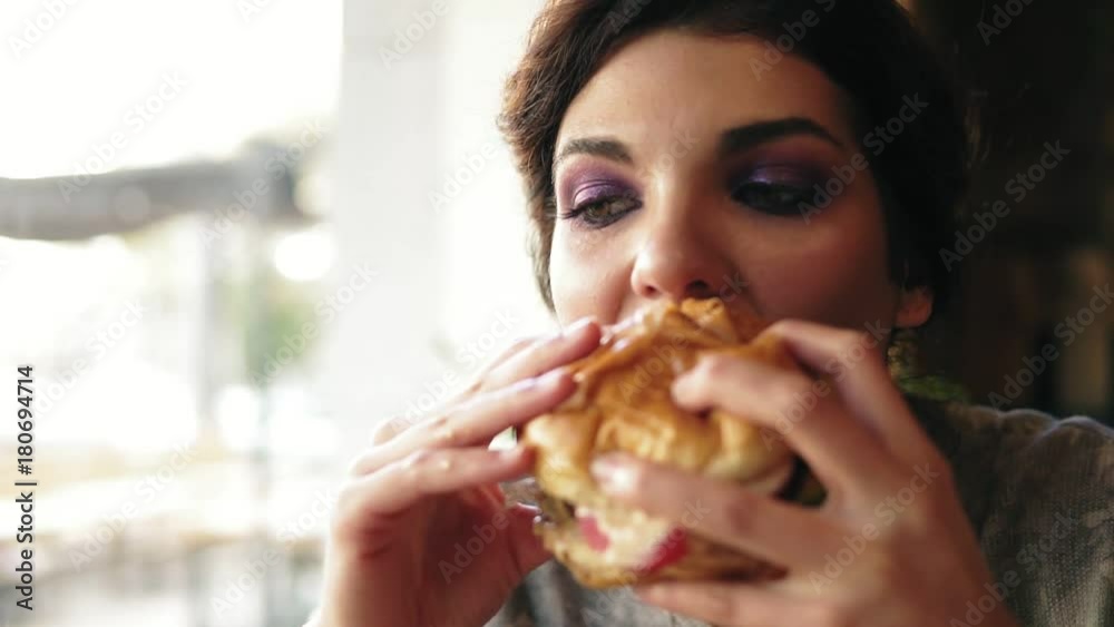 Closeup view of young woman biting big tasty burger in cafe. Slowmotion shot Closeup view of young woman biting big tasty burger in cafe. Slowmotion shot
