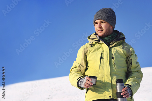 Man drinking hot coffee in thermos mug and looking into the mountains in snow, winter hike © Aleksey