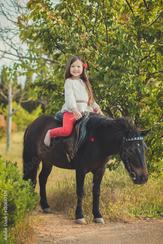 Girl with brunette hair and brown eyes stylish dressed wearing rustic village clothes white shirt and red pants on belt posing with sitting on black young horse pony © TwinkleStudio