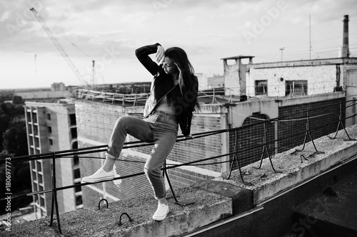 Portrait of a gorgeous young woman in black leather jacket, jeans and sneakers sitting on handrails on the roof with picturesque view of a park. Black and white photo.