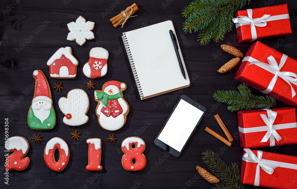 Christmas background with mobile phone with blank white screen, notebook with pen, ginger cookies, red gift boxes and fir tree branches on wooden background, copy space. Top view, flat lay
