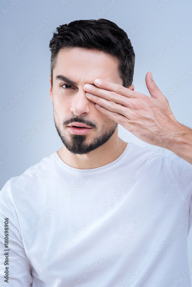 Poor eyesight. Calm attentive serious young man looking not glad while closing his right eye and realizing the bad condition of his eyesight Poor eyesight. Calm attentive serious young man looking not glad while closing his right eye and realizing the bad condition of his eyesight