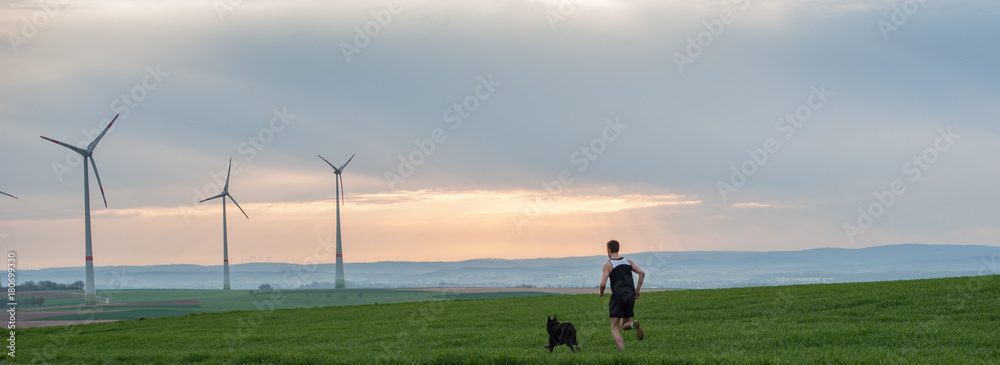 Obraz premium a man runs along the green field with a dog, with a view of the windmills