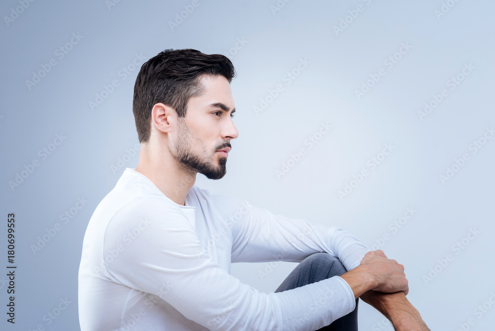 Peaceful mood. Pleasant handsome young man sitting quietly against the blue background while looking into the distance and thinking about his future Peaceful mood. Pleasant handsome young man sitting quietly against the blue background while looking into the distance and thinking about his future