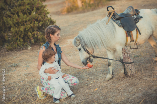 Romantic scene beautiful lady young mother with her cute baby daughter enjoy time together in city village park walking feeding white horse pony photo