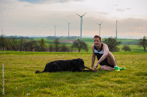 Young man sits on the field with a labrador, at sunset with a view of the windmills © Pavel
