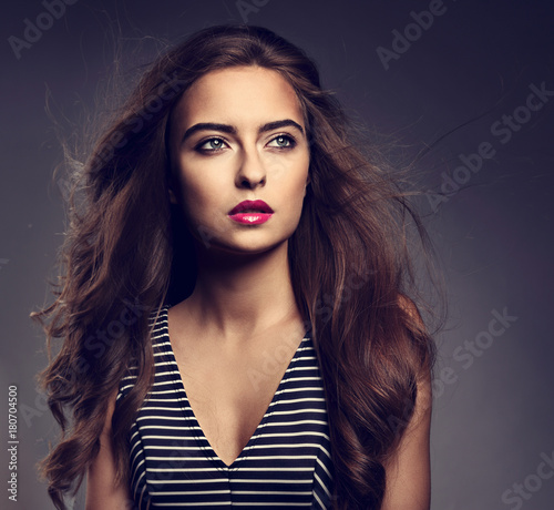 Beautiful woman with bright makeup and wind long hair, pink lipstick looking up in striped dress on grey background. Closeup toned bright portrait