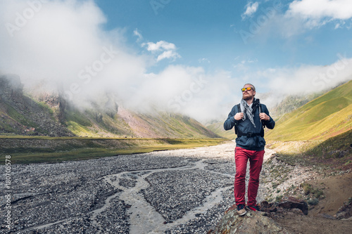 Young Male Traveler With Backpack Walks Along Valley Of A Mountain River Against The Background Of Mountains And Clouds