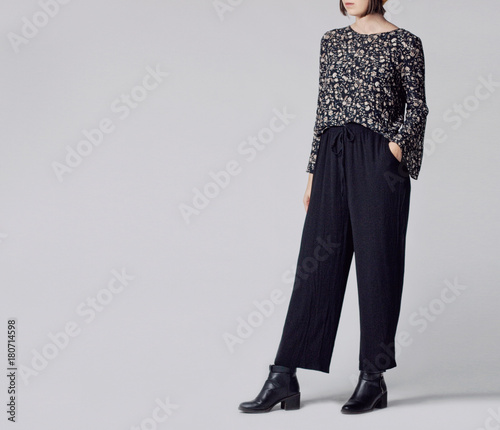 Foto Woman wearing stylish outfit with black patterned blouse, black high-waisted wide leg trousers and black ankle boots isolated on grey background