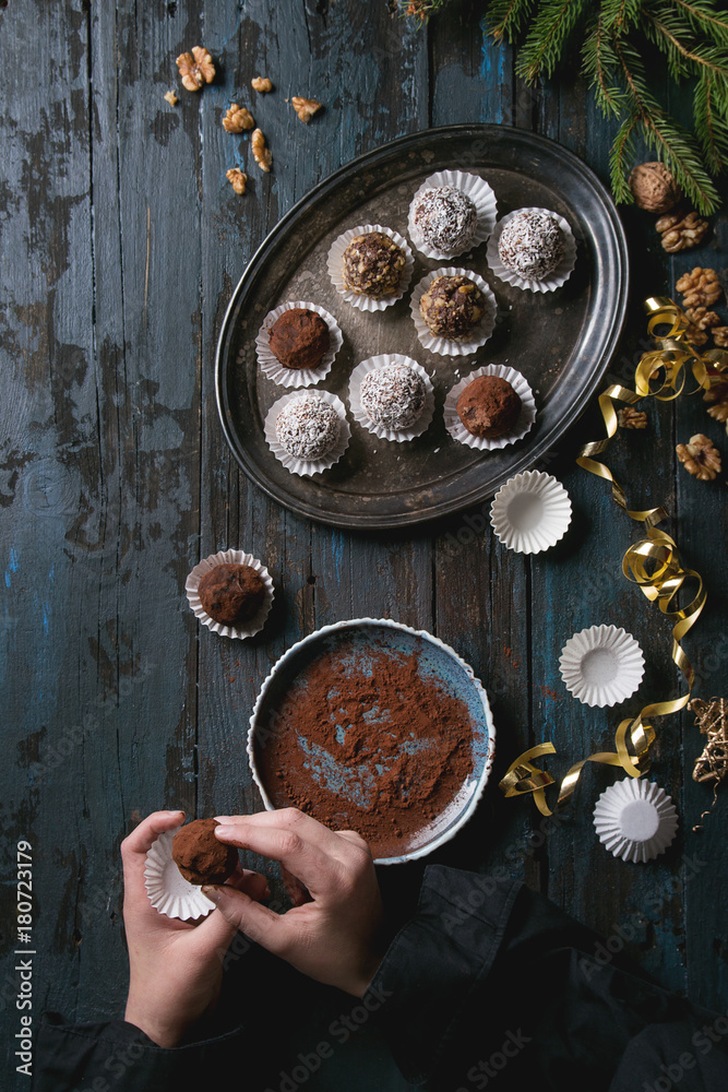 Child hands make homemade dark chocolate truffles with cocoa powder, coconut, walnuts as Christmas gift, put on vintage tray. Fir tree, Christmas decorations above on old wooden table. Top view. Child hands make homemade dark chocolate truffles with cocoa powder, coconut, walnuts as Christmas gift, put on vintage tray. Fir tree, Christmas decorations above on old wooden table. Top view.