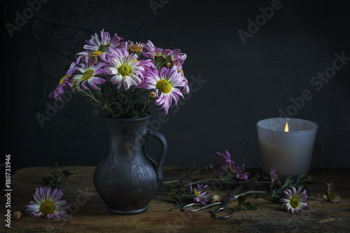 .Still life with autumn chrysanthemums in a vase, candles and old scissors on a dark rustic wooden table and dark background.  Low key. Focus concept.
