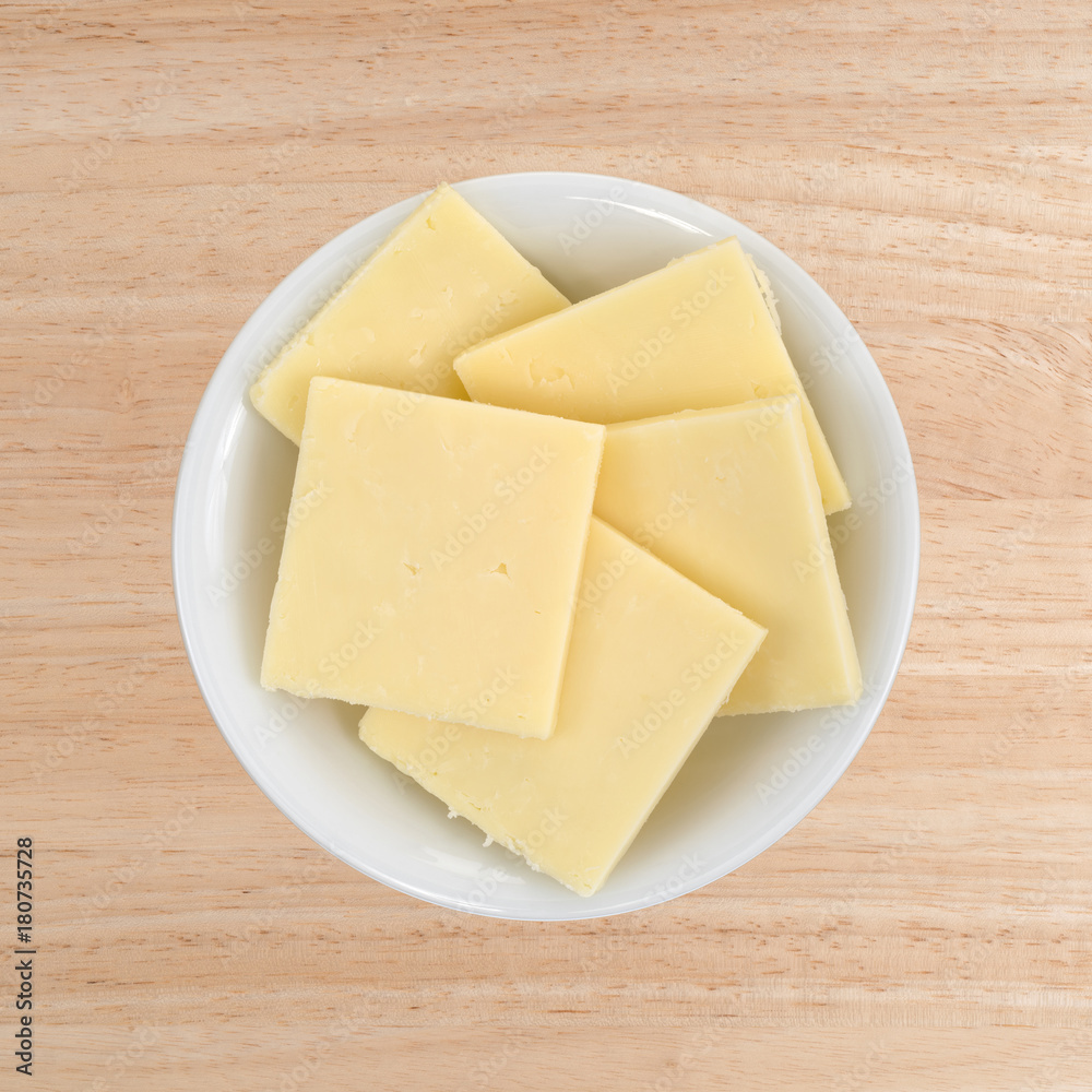 Top view of a small white bowl filled with sharp cheddar cheese square slices on a wood table. Top view of a small white bowl filled with sharp cheddar cheese square slices on a wood table.