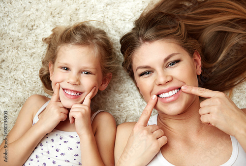 Portrait of beautiful daughter with mom showing their healthy white smile lying on carpet at home.
