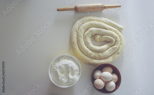 on a white table a bowl with eggs and flour, ready to be rolled up and next to the rolling pin. view from above