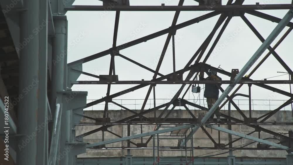 The welder in the protective helmet works at a height, welding the frame elements. Construction work: the creation of the roof of the premises, the metal frame of the building. Welder at work. The welder in the protective helmet works at a height, welding the frame elements. Construction work: the creation of the roof of the premises, the metal frame of the building. Welder at work.