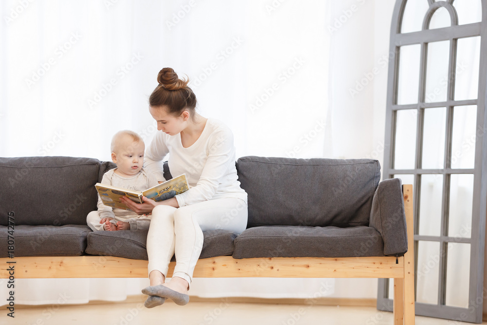 Pretty young mother reading a book to her little baby boy on a sofa in a light bedroom of house. Family holiday and togetherness.