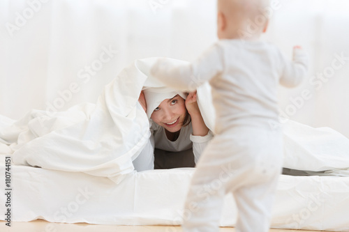 happy young mother plays at hide-and-seek with her baby boy in a bed in a light bedroom of the house. Mother's Day. White pajamas, happy childhood.