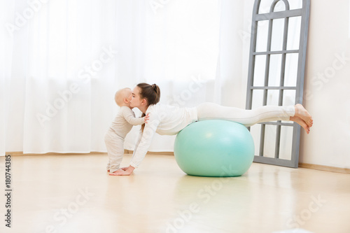 Happy young mother with baby wearing white sport clothes doing gymnastics with green Exercise ball and having fun at light domestic home interior.