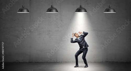 Obraz na plátně A scared businessman stands cowering under the rays of industrial pendant lights right above him