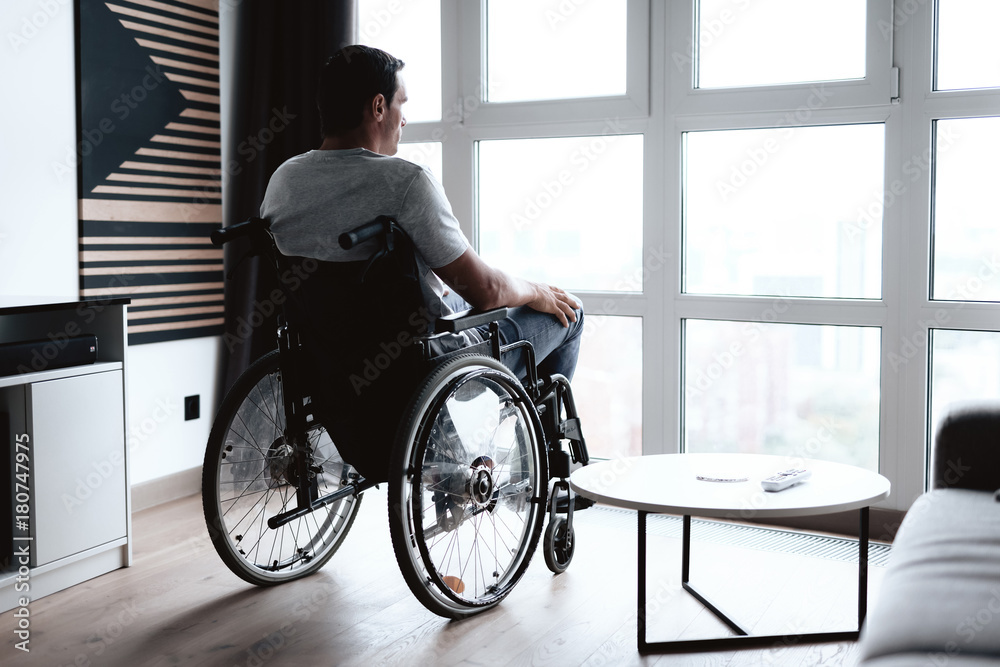 The disabled person in a wheelchair sits in front of a large panoramic window and looks into it. He is sad. He is in the living room of his modern apartment. The disabled person in a wheelchair sits in front of a large panoramic window and looks into it. He is sad. He is in the living room of his modern apartment.
