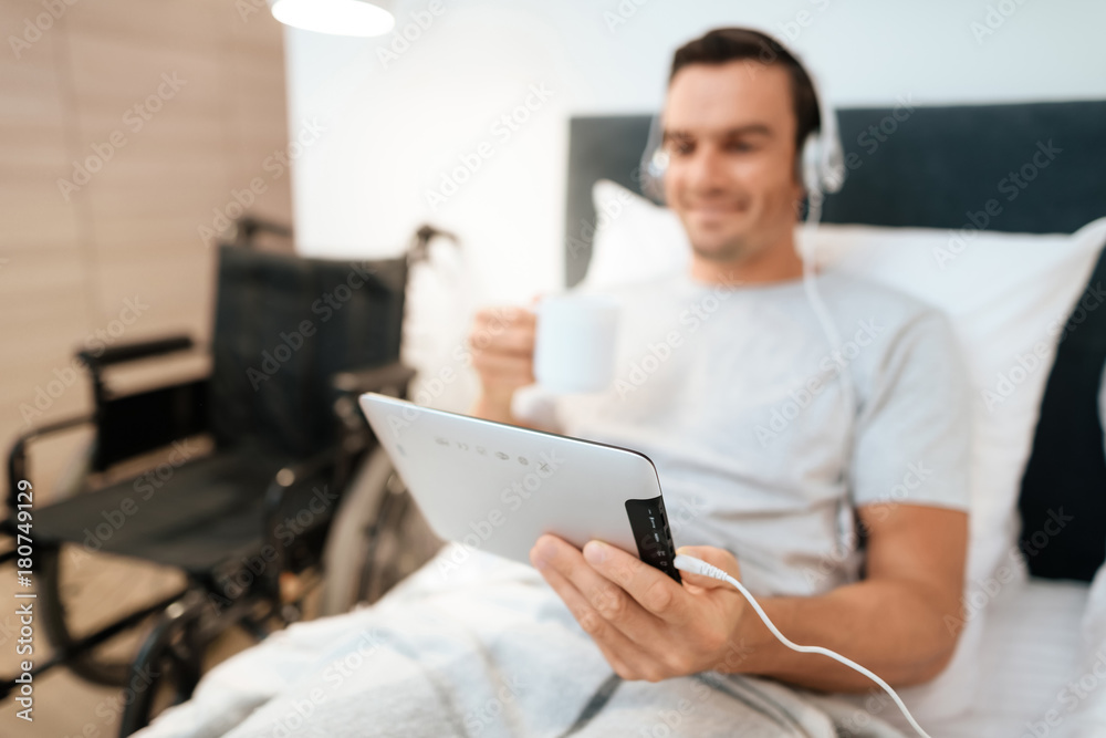 The invalid lies in bed and looks something on the tablet. He smiles. because he is happy. Next to his bed is his black wheelchair. It lies on a large white step.