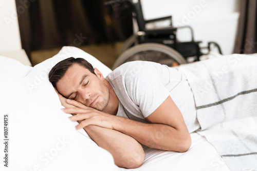 The disabled person sleeps in his white bed. He's sleeping. Behind him is his wheelchair. He is in the bedroom of his modern apartment. The man is sleeping in a white T-shirt.