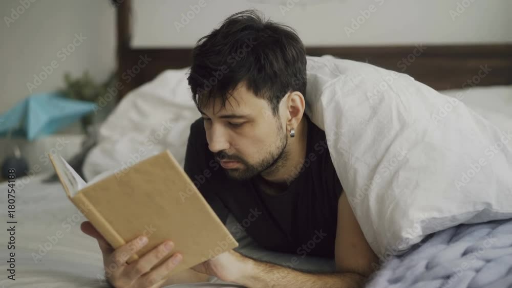 Handsome young man reading a book lying in bed under blanket at home Handsome young man reading a book lying in bed under blanket at home