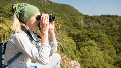 A blond Woman tourist with a backpack on a cliff looking through binoculars on the sea. Travel concept.