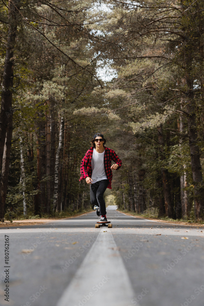 A young hipster in a cap and plaid shirt is riding his longboard on a country road in the forest A young hipster in a cap and plaid shirt is riding his longboard on a country road in the forest