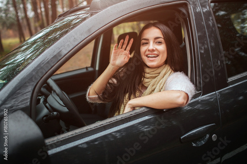 Long-haired brunette on the auto background. A female model is wearing a sweater and a scarf. Autumn concept. Autumn forest journey by car