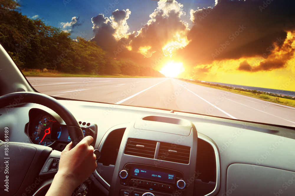 Car dashboard with driver's hand on the steering wheel against the empty road and sky with sunset