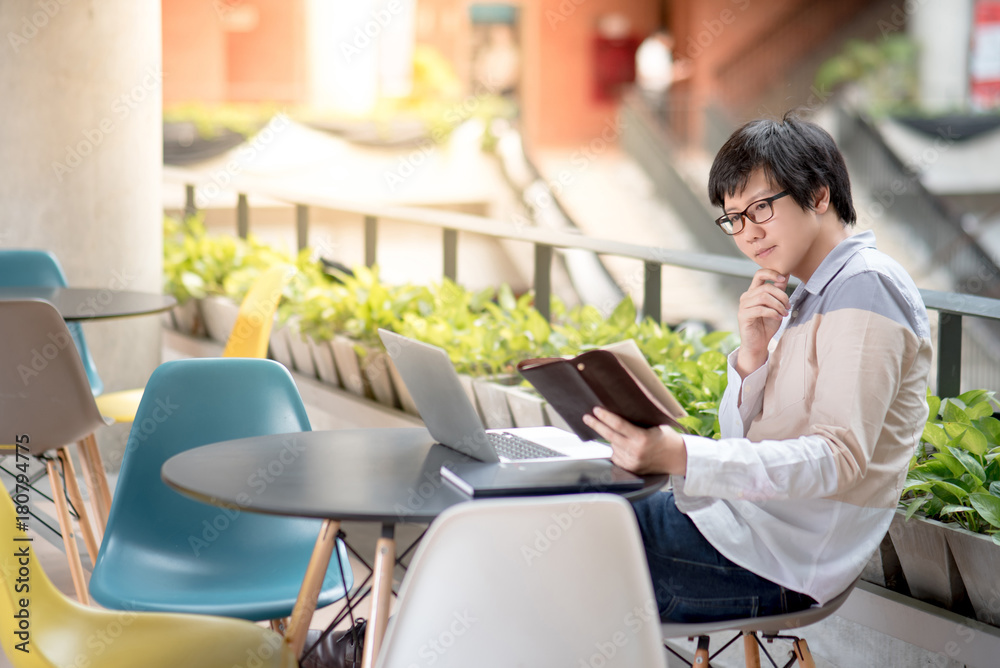 Young Asian student man dressed in casual style reading book and using laptop computer in college building, education research and self learning in university life concepts