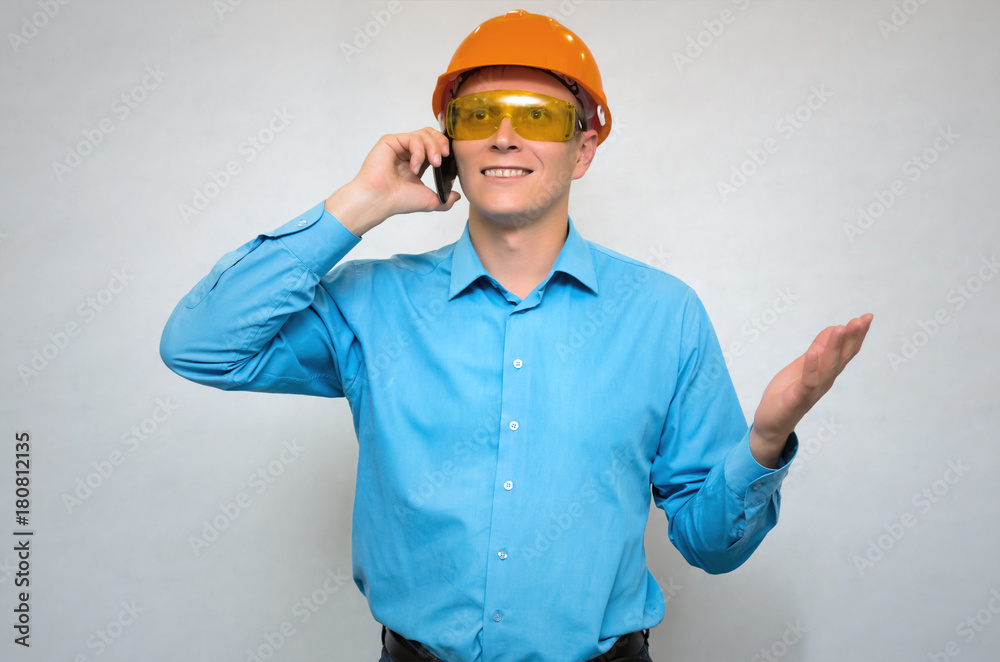 Young builder worker in hardhat helmet speaks by mobile phone, gestures by hand and looking up isolated.