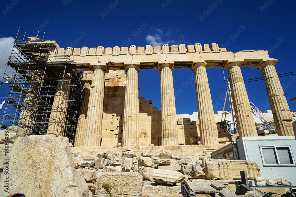 Fototapeta premium Restoration work in progress at world heritage classical Parthenon on marble block base on top of Acropolis with site office, scaffolding and blue sky background