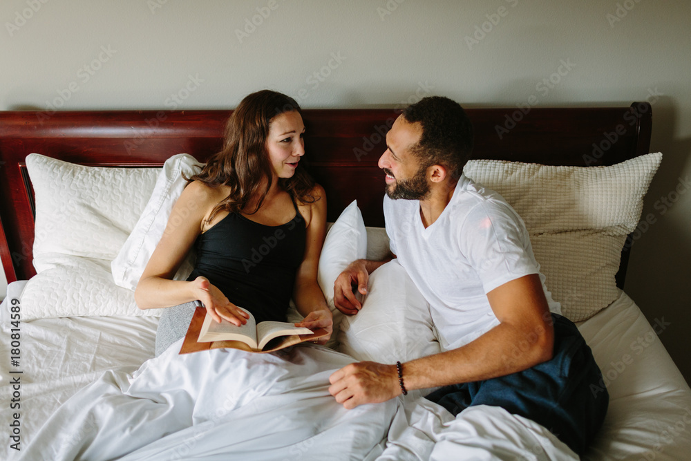 Happy mixed race couple in bed talking and reading a book